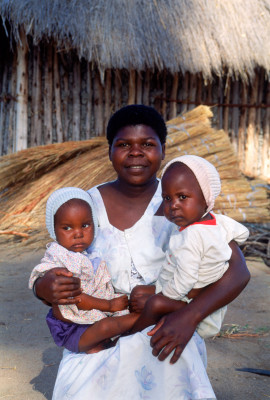 African woman holding two babies in her arms in front of village hut in rural Zimbabwe