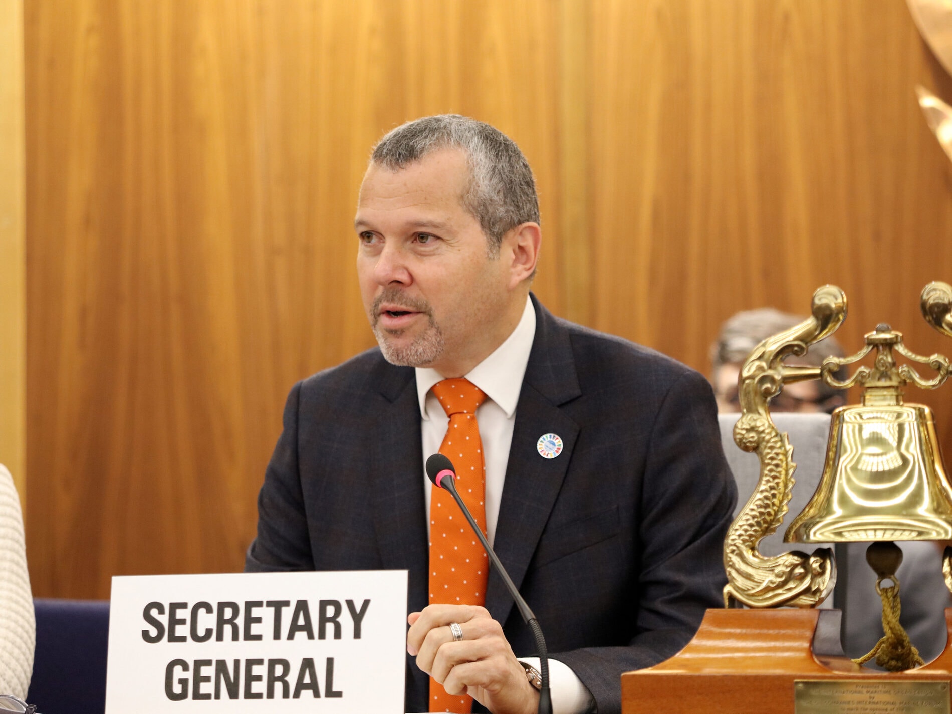 A man speaks into a microphone while sitting behind a sign reading "Secretary General."