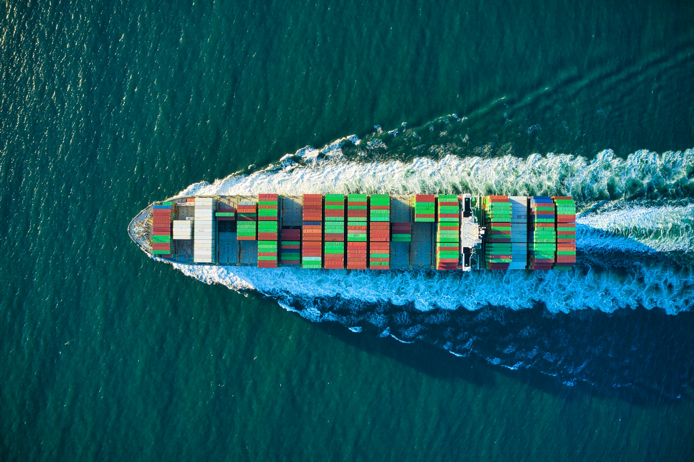 A cargo ship covered with containers leaves a wake as it passes over the surface of the ocean.