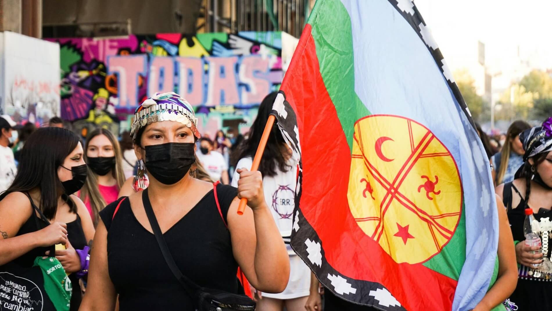 A woman wearing a bandana and black surgical mask carries a flag.