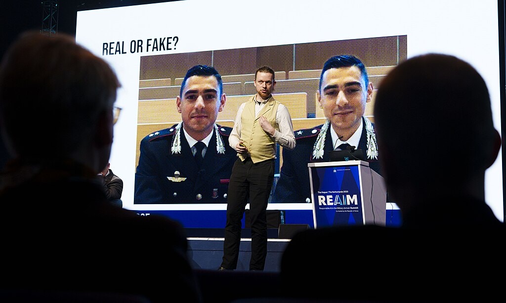 REAIM speaker stands in front of an image that reads "Real or fake?"