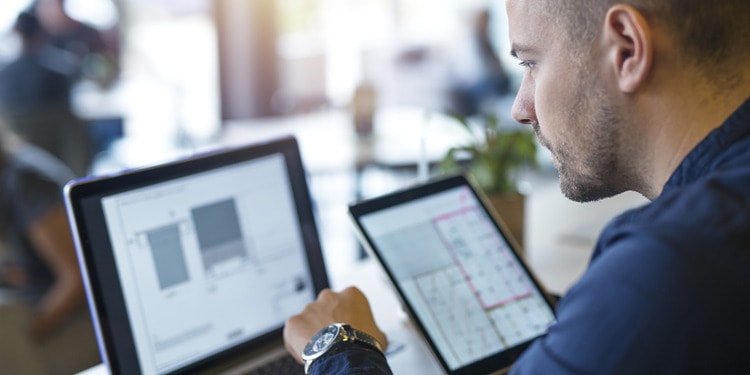 Team member of a logistics software development services company looking at his screen with a tablet in his right hand and typing with his left hand.
